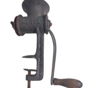 May include: An antique Regal meat grinder made of dark metal. The grinder has a hand crank with a wooden handle and a clamp to secure it to a surface. The word "REGAL" is embossed on the grinder's body.