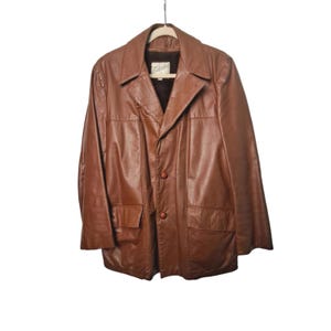 May include: A brown leather jacket with a collared neckline, two front pockets, and three buttons. The jacket is hanging on a wooden hanger against a white background. The jacket appears to be vintage.