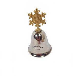 May include: A silver bell with a gold snowflake topper. The bell has the word "Christmas" and the year "1994" engraved on it. The bell is a decorative item, likely for the holiday season.