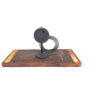 May include: An antique clock escapement mechanism, featuring a dark metal balance wheel and a circular dial. The mechanism is mounted on a rectangular wooden base with a distressed finish, showcasing a vintage aesthetic.