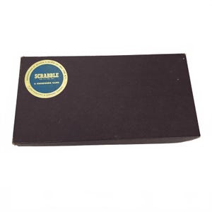 May include: A rectangular, dark brown Scrabble game box. A circular sticker on the top of the box reads "SCRABBLE A CROSSWORD GAME". The sticker is blue and white with gold lettering. The box is closed and appears to be vintage.