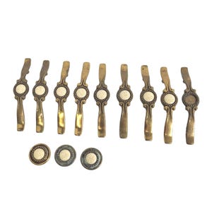May include: A collection of antique brass drawer pulls and matching knobs. The pulls have a decorative design with a central cream-colored insert. The knobs also feature the same cream-colored insert.