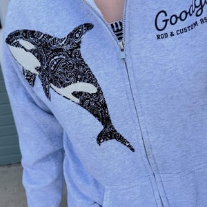 May include: A light gray zip-up hoodie featuring a black and white orca graphic with a detailed paisley pattern on the chest. The text Goodguys ROD & CUSTOM ASSOCIATION is printed in black on the upper right side near the silver metal zipper.