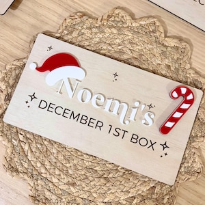 May include: A rectangular light-coloured wooden sign featuring the white text Noemi's and the black text DECEMBER 1ST BOX. The sign is decorated with a red and white acrylic Santa hat, a red and white striped candy cane, and small star icons.