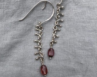 Könnte beinhalten: Ein Paar silberne Hängeohrringe. Jeder Ohrring hat ein kettenartiges Design mit kleinen, gebogenen Akzenten und einem tropfenförmigen, rot-violetten Edelstein am unteren Ende. Die Ohrringe hängen an gebogenen Ohrhaken.