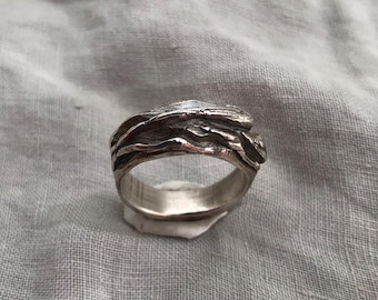 May include: A silver ring with a textured, organic design. The ring features a layered, flowing pattern, creating a unique and artistic look. The ring is displayed on a neutral gray surface, highlighting its metallic sheen.