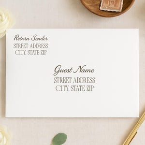 May include: A white envelope with elegant script text for return and guest addresses. The scene includes a gold pen, paperclip, and floral accents, suggesting a wedding or event invitation theme.