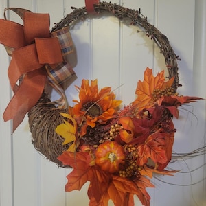 May include: A fall-themed wreath with a brown twig base, decorated with a red and white plaid bow, a small orange pumpkin, a pine cone, and orange, red, and yellow fall leaves.