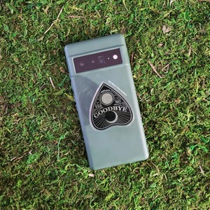 May include: A green phone case with a black and white ouija board sticker that says "Hello, Yes, Goodbye, No".