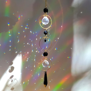 Peut inclure: Un attrape-soleil suspendu avec des anneaux dorés, des sphères de cristal transparentes et des perles noires. Un cristal noir pointu est suspendu en bas. Le capteur solaire est suspendu à une chaîne dorée. Des motifs lumineux arc-en-ciel sont projetés sur le fond.