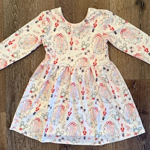 Puede incluir: Vestido blanco de manga larga con un estampado festivo con cascanueces, lazos y coronas con forma de Mickey Mouse. El vestido tiene un escote redondo y una falda acampanada, perfecto para una celebración.
