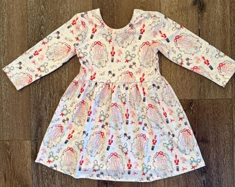 May include: A white long-sleeved dress with a festive pattern featuring nutcrackers, bows, and Mickey Mouse-shaped wreaths. The dress has a scoop neckline and a flared skirt, perfect for a holiday celebration.
