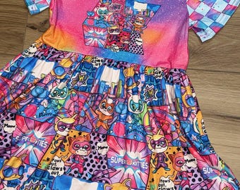 May include: A colorful dress with a V-neck and short sleeves. The bodice features a vibrant tie-dye design with a graphic of cartoon cats. The skirt is patterned with comic book style illustrations of superhero cats, with text that says "Super Kitties".