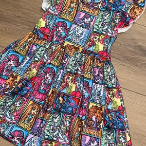 Op de afbeelding: Een kleurrijke jurk met korte mouwen en een levendige print van stripfiguren in een patchwork-ontwerp. De jurk heeft een witte halslijn en fladderende mouwen. De print bevat de namen "CLEO" en "CLAWDEEN".