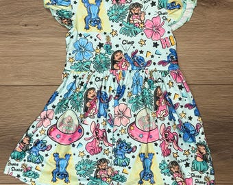 May include: A light blue dress with a colorful print featuring cartoon characters, flowers, and stars. The dress has short, ruffled sleeves and a round neckline. The print includes the words "Clap", "Shake", and "Wave".