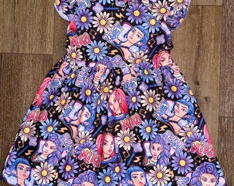 May include: A black dress with a vibrant print featuring stylized portraits, daisies, and text. The dress has a round neckline, short flutter sleeves, and a gathered waist. The text includes the name "Mira".