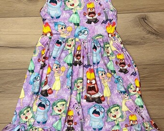 May include: A sleeveless purple dress featuring a repeating pattern of animated characters. The dress has a ruffled hem and a scoop neckline with yellow trim. The characters are in various poses and expressions.