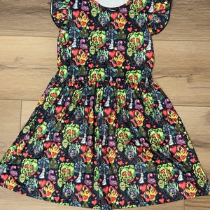 May include: A black dress with a colorful pattern featuring green poison apples, red hearts, and other whimsical designs. The dress has short sleeves and a gathered waist, creating a flared skirt. The fabric has a white neckline.