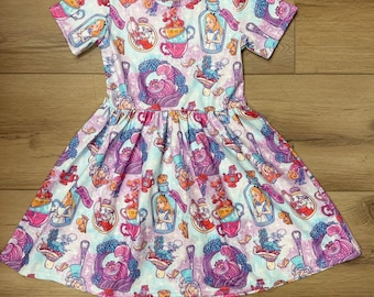 May include: A short-sleeved dress featuring a colorful pattern of Alice in Wonderland characters and elements. The design includes Alice, teacups, bottles, and the Cheshire Cat in shades of pink, purple, blue, and white. The dress has a gathered skirt and a scoop neckline.