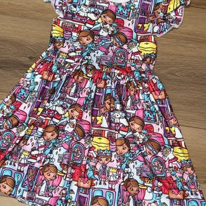 May include: A colorful, short-sleeved dress featuring a repeating pattern of cartoon characters and various objects in shades of pink, purple, blue, and yellow. The dress has a round neckline and ruffled sleeves, with a gathered waist and a flared skirt.