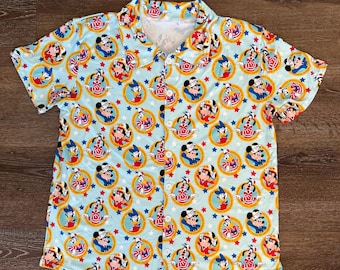 May include: Light blue button-down shirt with a repeating pattern of cartoon characters in circular frames. The characters include Mickey Mouse, Donald Duck, and others, set against a background of stars.
