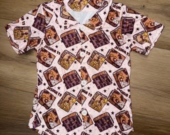 May include: A short-sleeved button-down shirt with a light pink background. The shirt is patterned with brown and tan books, stars, and the text "JOSHUA 1:9". The shirt has white buttons and a collar.