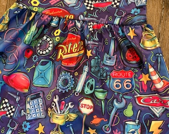 May include: A blue dress featuring a colorful pattern of automotive-themed graphics. The design includes images of tools, racing flags, Route 66 signs, and the word "Cars". The dress has short sleeves and a gathered waist.