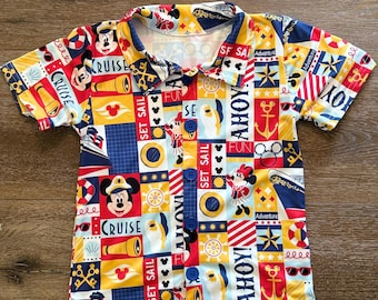 May include: A short-sleeved button-down shirt with a colorful nautical-themed print. The shirt features Mickey Mouse, anchors, and the words "Cruise" and "Ahoy!" in a patchwork design. The shirt has blue buttons and a collar.