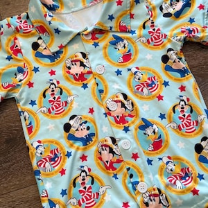 May include: Light blue button-up shirt featuring a repeating pattern of Mickey Mouse, Donald Duck, and Goofy in sailor outfits, surrounded by gold circles, red and blue stars. The shirt has a collared neckline and white buttons.