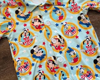 May include: Light blue button-up shirt featuring a repeating pattern of Mickey Mouse, Donald Duck, and Goofy in sailor outfits, surrounded by gold circles, red and blue stars. The shirt has a collared neckline and white buttons.