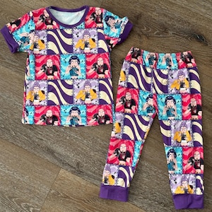 May include: A two-piece pajama set featuring a short-sleeved top and matching pants. The set is covered in a colorful print of cartoon characters in squares, with purple and yellow wavy stripes. The cuffs and neckline are purple.