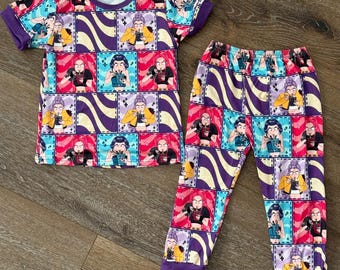 May include: A two-piece pajama set featuring a short-sleeved top and matching pants. The set is covered in a colorful print of cartoon characters in squares, with purple and yellow wavy stripes. The cuffs and neckline are purple.