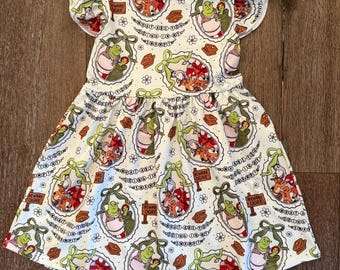 May include: White cotton dress with a repeating pattern of cartoon characters in oval frames, bows, and floral accents. The dress has short, ruffled sleeves and the text "BEWARE OGRE" on some of the frames.