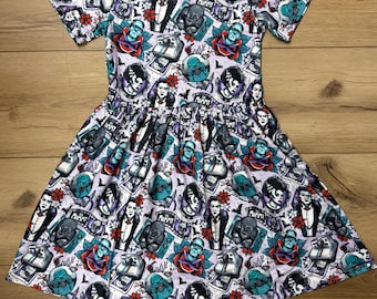 May include: A short-sleeved dress with a light purple background and a repeating pattern of classic monster movie characters. The dress features a gathered waist and a round neckline. The print includes images of Frankenstein's monster, Dracula, and the Mummy.