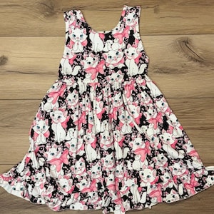 May include: A sleeveless dress featuring a repeating pattern of white cartoon cats with pink bows on a black background. The dress has a gathered waist and a flared skirt, suitable for children.