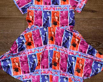 May include: A short-sleeved dress with a vibrant pattern featuring a repeating design of a woman in a red jacket and the text "INK HUNTRX". The dress has a scoop neckline and a flared skirt, with a color palette of pink, purple, blue, and orange.