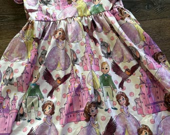 May include: A white dress with a pattern featuring cartoon characters, castles, birds, and polka dots in shades of pink, purple, and green. The dress has short sleeves and a gathered waist, suitable for a child.