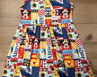 May include: A sleeveless dress with a nautical theme. The dress features a colorful patchwork design with images of Mickey and Minnie Mouse, cruise ships, anchors, and the words "CRUISE", "AHOY", and "SET SAIL". The dress is made of a lightweight fabric.