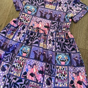 Może przedstawiać: Fioletowa sukienka z nadrukiem w stylu komiksowym z ilustracjami postaci, tekstem i wzorami gwiazd. Sukienka ma krótkie rękawy i rozkloszowaną spódnicę. Tekst zawiera zwroty takie jak "Take Down" i "Gonna Be Golden".