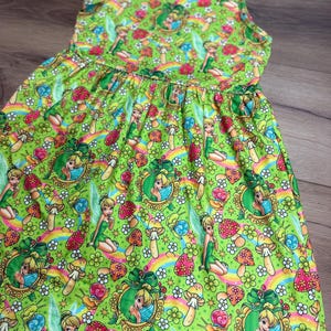 May include: A sleeveless green dress featuring a repeating pattern of a fairy in a gold frame, mushrooms, rainbows, flowers, and snails. The dress has a gathered waist and a flared skirt. The fabric is a bright, vibrant green.