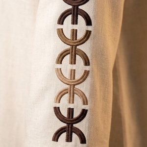 May include: Close-up of a beige linen garment sleeve. The sleeve features an embroidered design in shades of brown. The design consists of repeating, stylized geometric shapes. The fabric has a textured appearance.
