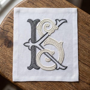 Puede incluir: Una servilleta de tela blanca cuadrada con un monograma bordado de las letras K y S. La letra K está cosida con hilo gris oscuro, mientras que la letra S está entrelazada con hilo color crema, ambos con detalles decorativos de tipo serif.