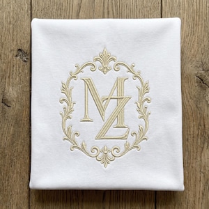 May include: A square white fabric item featuring centered embroidery in a light beige thread. The design consists of the letters M and Z intertwined, enclosed within an ornate, symmetrical floral frame with decorative scrollwork and fleur-de-lis accents.