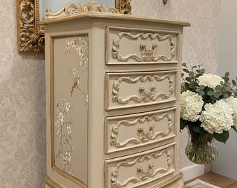 May include: A tall, cream-colored chest of drawers featuring four drawers with ornate, raised molding and gold-toned hardware. The side panel displays a delicate botanical illustration of birds and flowering branches in soft, muted tones.