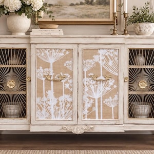 May include: A cream-colored wooden sideboard featuring two central doors with white botanical floral prints on a tan background and two side doors with gold wire sunburst patterns. The piece includes ornate gold handles and carved detailing.