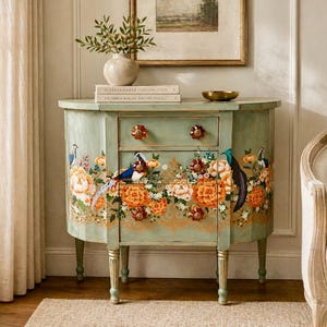 May include: A mint green painted cabinet with two drawers and a lower cabinet door. The cabinet is decorated with orange and yellow floral patterns and images of birds. The cabinet has a curved front and gold-tipped legs.