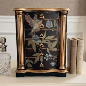 May include: A black jewelry chest with gold trim and a bird and floral design. The chest has five drawers with gold handles. A clear glass decanter and two books are on the table.