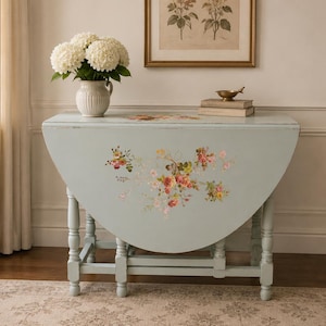 May include: A light blue drop-leaf table with a painted floral motif featuring pink, yellow, and green flowers on the front panel. The table has turned wooden legs and a distressed finish, with a white pitcher of white hydrangeas on top.