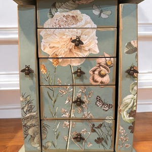 May include: A decorative jewelry box with a light blue finish and gold trim. The box features floral and butterfly designs on the drawers and sides, with bee-shaped drawer pulls. The drawers are adorned with large flower illustrations.