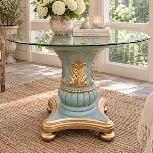 May include: A round glass-top table featuring a pedestal base painted in a distressed light blue finish with ornate gold-toned carved detailing. The base has a square-shaped bottom with four rounded feet and a central column with fluted accents.